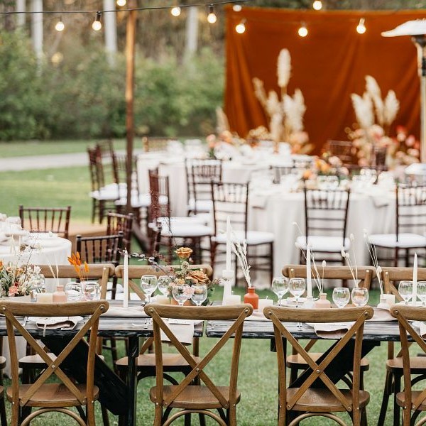 Organization of stylish wedding and new normal at outdoor in summer. Candles, flowers, accessories, glasses and plates on table for guests, wooden chairs on green lawn with photo zone with dry plants