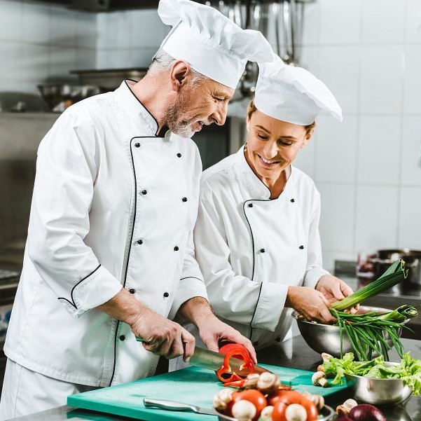 smiling female and male chefs in uniform and hats cooking in restaurant kitchen